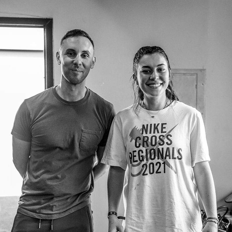 A man and a woman stand side by side, smiling. The woman is wearing a "Nike Cross Regionals 2021" T-shirt. The image is in black and white and appears to be taken indoors.