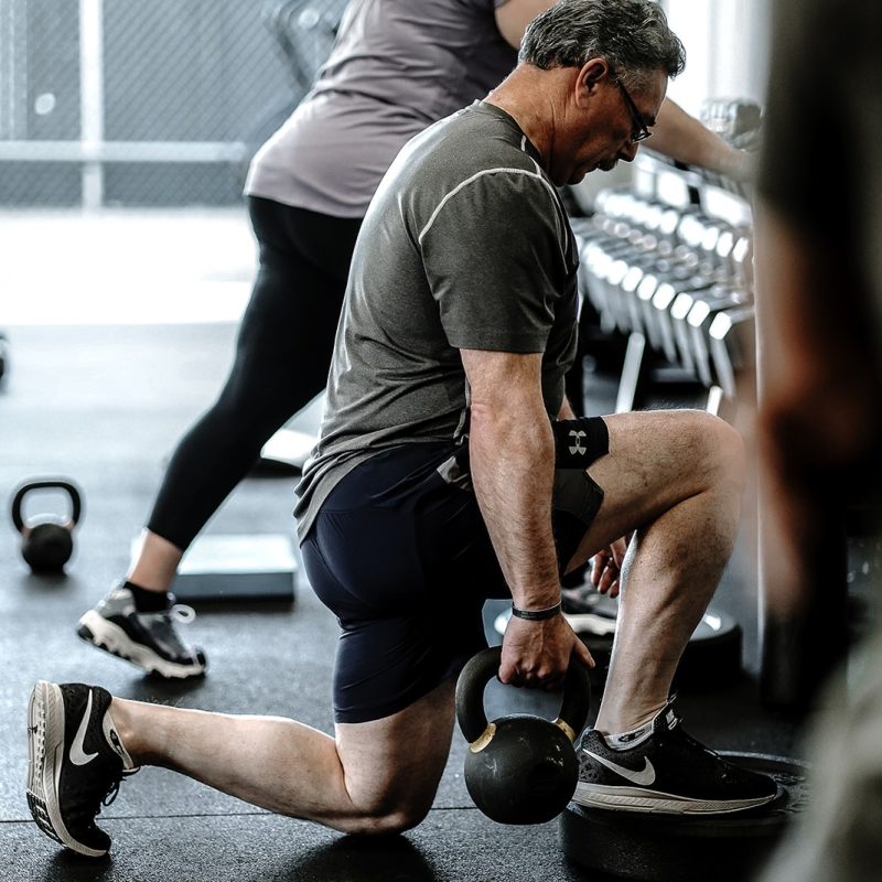 A man lunges while holding a kettlebell in one hand at a gym, with a woman in the background reaching for a dumbbell on a rack. Both are dressed in workout clothes.