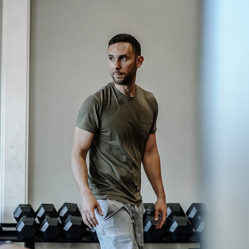 A man in a khaki t-shirt and light gray pants stands in a gym, looking to the side, with a row of black dumbbells visible behind him.