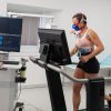 A woman runs on a treadmill wearing fitness sensors and a breathing mask while a healthcare professional monitors data on multiple screens in a medical testing room.