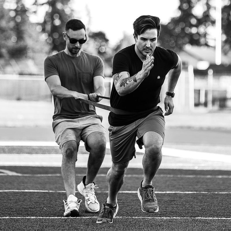 A man sprints forward on a field with determination while another man behind him holds a resistance band around his waist, providing resistance during the workout. Both are wearing athletic clothing. The image is in black and white.