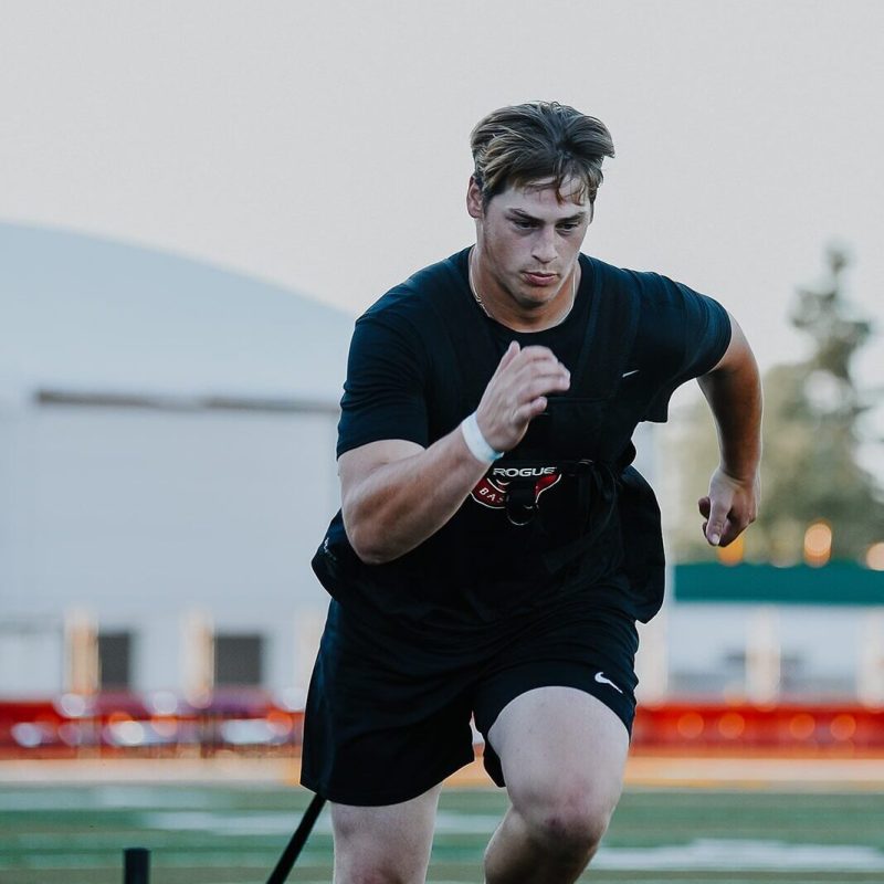 A young man wearing athletic gear sprints forward on a field while pulling a resistance sled, focused on his workout. The background features a blurred sports facility and green trees.